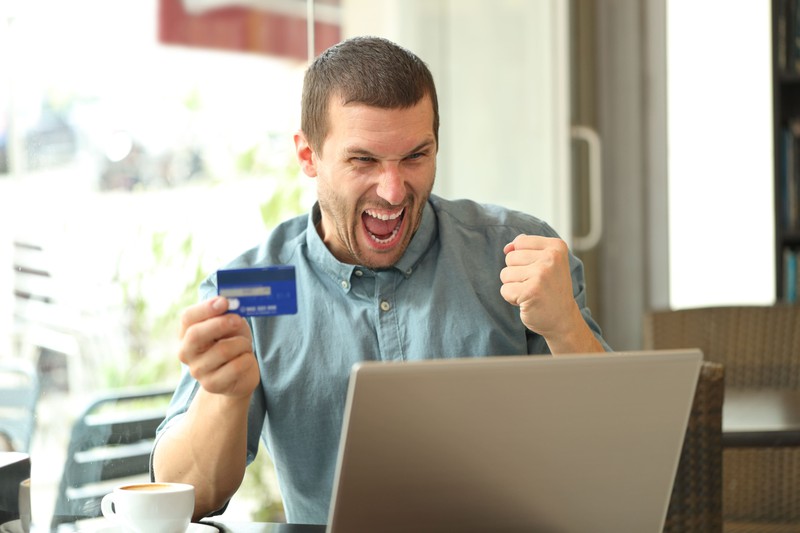 Ein fröhlicher Mann sitzt vorm Laptop und hält eine Kreditkarte in der Hand.