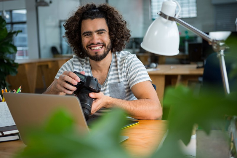 Ein junger Mann sitzt vor einem Laptop und hält eine Kamera in der Hand.