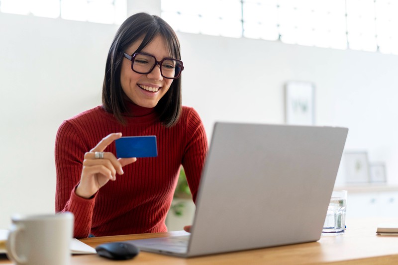 Zu sehen ist eine Frau, die am Laptop sitzt und eine Kreditkarte in der Hand hält.