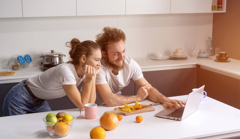 Zu sehen sind ein Mann und eine Frau, die in der Küche stehen und auf einen Laptop schauen.