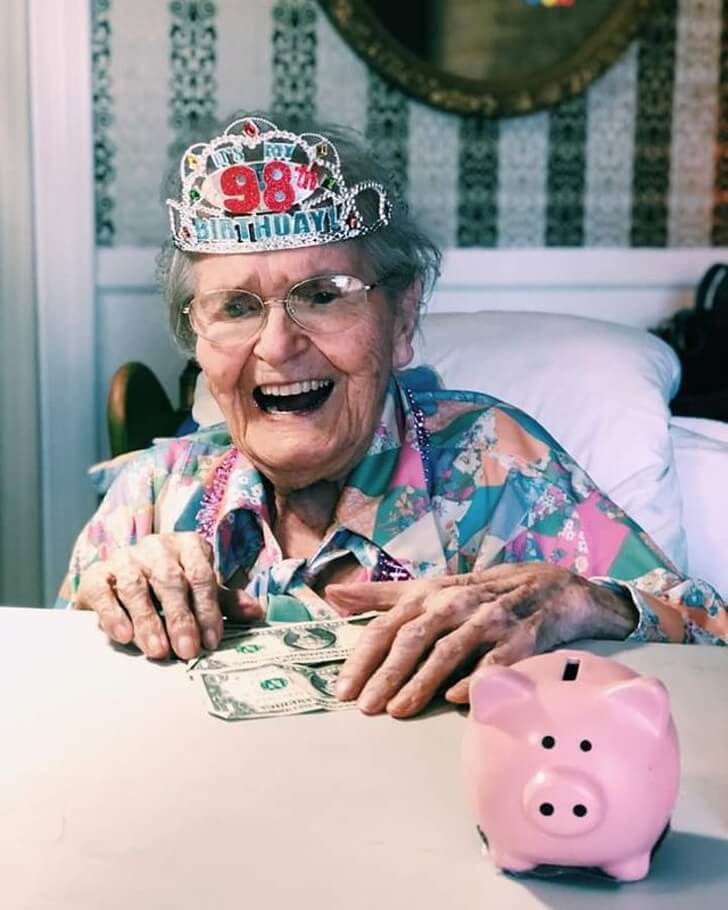 Urgroßmutter sitzt an ihrem 98 Geburtstag mit einer Krone und Kleingeld am Tisch und steckt es in ein Sparschwein