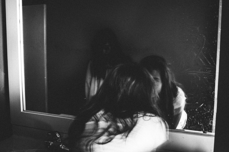 Zu sehen ist eine junge Frau, die in einen Spiegel schaut und es geht um Horror-Geschichten.