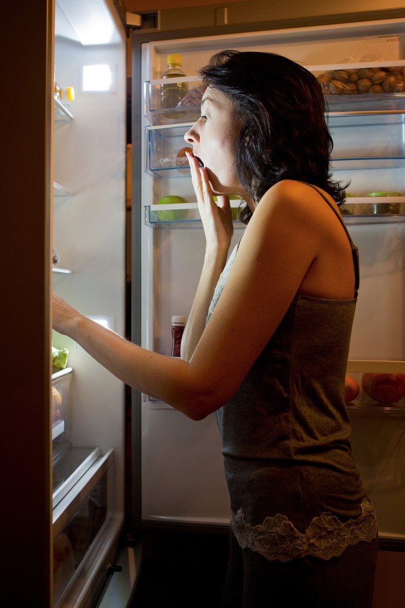Die müde Frau blick in den Kühlschrank, in den sie ihren Schlafanzug zum Kühlen gelegt hat.